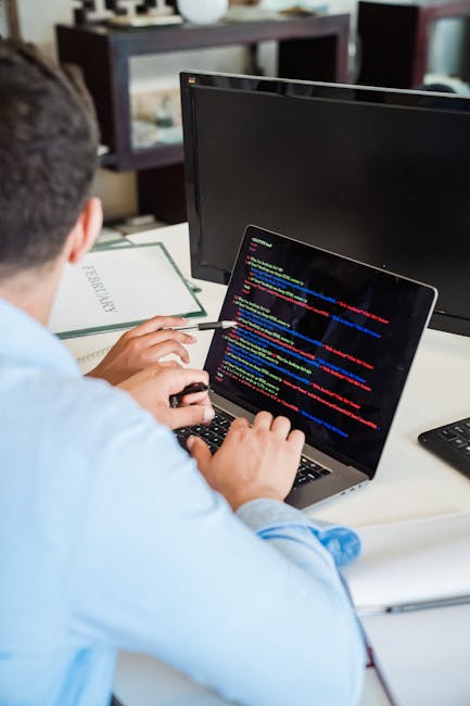 A person is seated at a desk working on a laptop displaying lines of colorful code, with a second monitor nearby. The workspace includes a book titled 'FREELANCER' partially visible next to the laptop